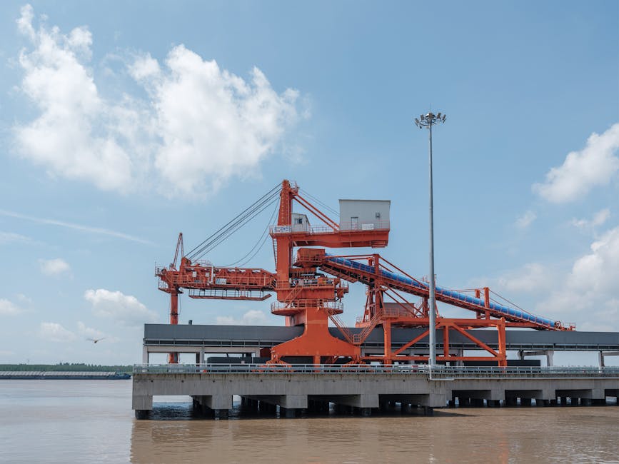 A large orange industrial crane situated at a waterside port under a blue sky.